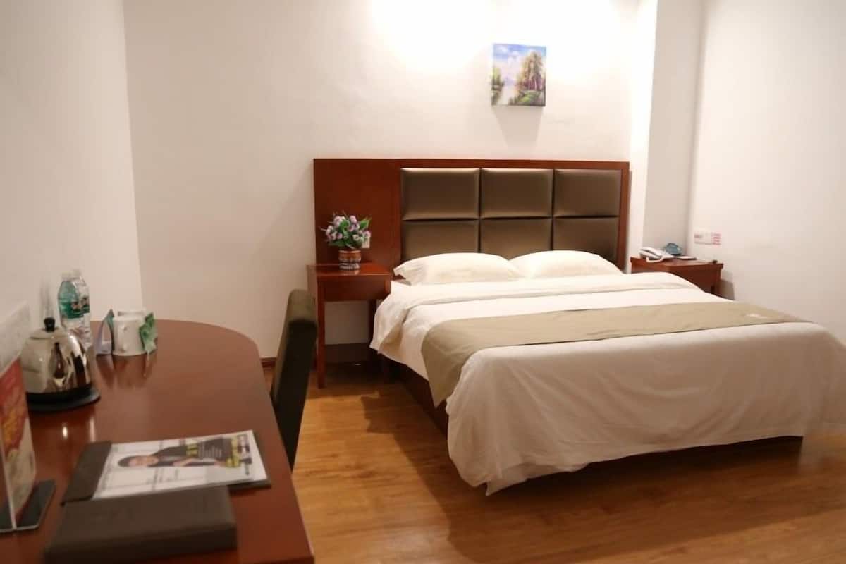 Une chambre d’hôtel avec un bureau en bois, un lit recouvert de draps blancs, une petite table de chevet ornée d’une plante et un tableau accroché au mur.