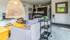 Un salon moderne avec un canapé gris, des coussins jaunes, un luminaire suspendu en osier et une cuisine en arrière-plan.