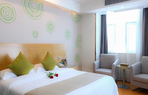 Ein Hotelzimmer mit einem Bett, zwei grünen Kissen, einem Nachttisch mit Lampe, einem Sessel und einem kleinen Tisch mit einem Buch.