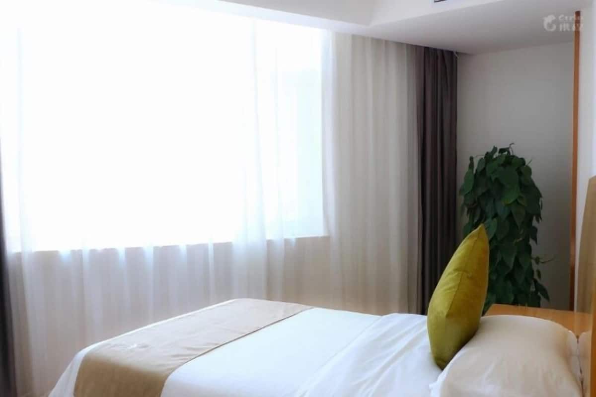 Een hotelkamer met een bed, een groot raam met dunne gordijnen, een potplant en een houten hoofdeinde.
