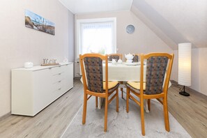 Un coin repas avec un meuble blanc, deux chaises en bois dotées d’assises rembourrées, une table dressée avec une théière, des tasses et une assiette.