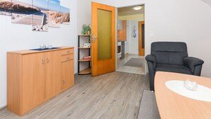 A room with a wooden cabinet, a black chair, a wooden table, and a door leading to a kitchen. - Vacation apartment 2 - House Wiederkehr in Bensersiel (Esens)