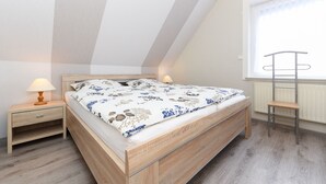 A bedroom with a wooden bed, two bedside tables with lamps, a radiator, and a window with a view. - Vacation apartment 2 - House Wiederkehr in Bensersiel (Esens)