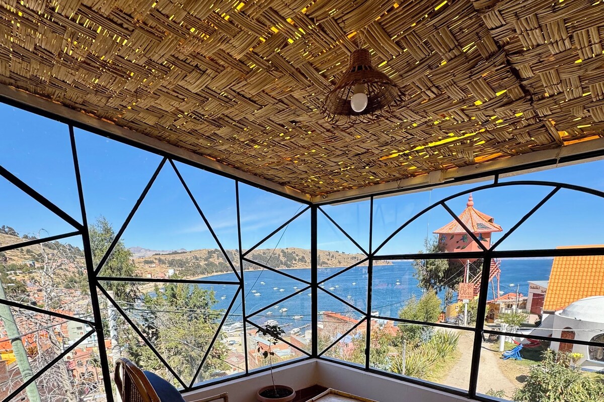 A room with a large window offering a view of a coastal town and the sea. There is a wicker chair with blue cushions and a small table.