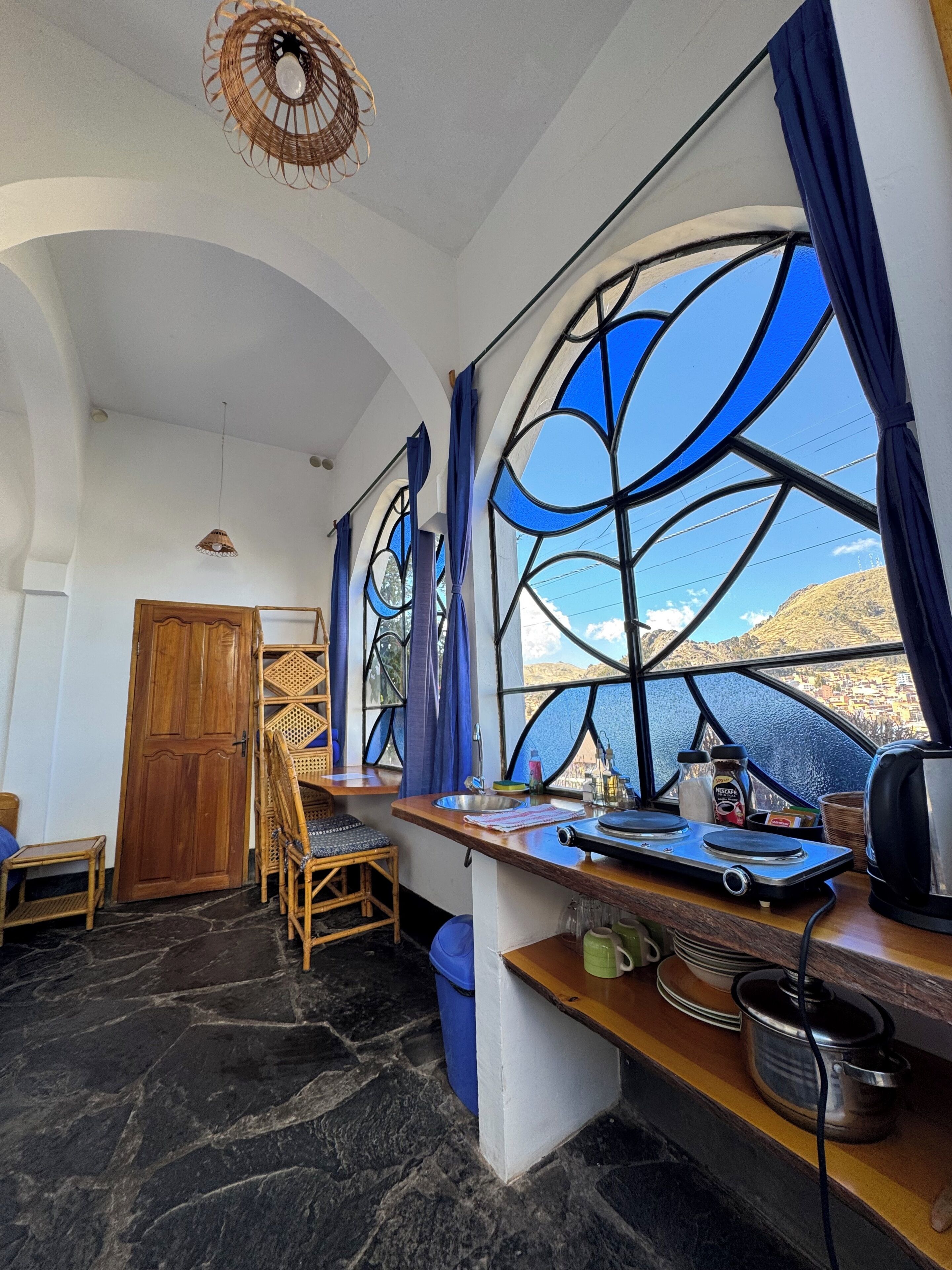 A room with a large window, a wooden door, a dining table with chairs, a sink, and a kettle.