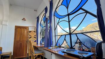 A room with a large window, a wooden door, a dining table with chairs, a sink, and a kettle.