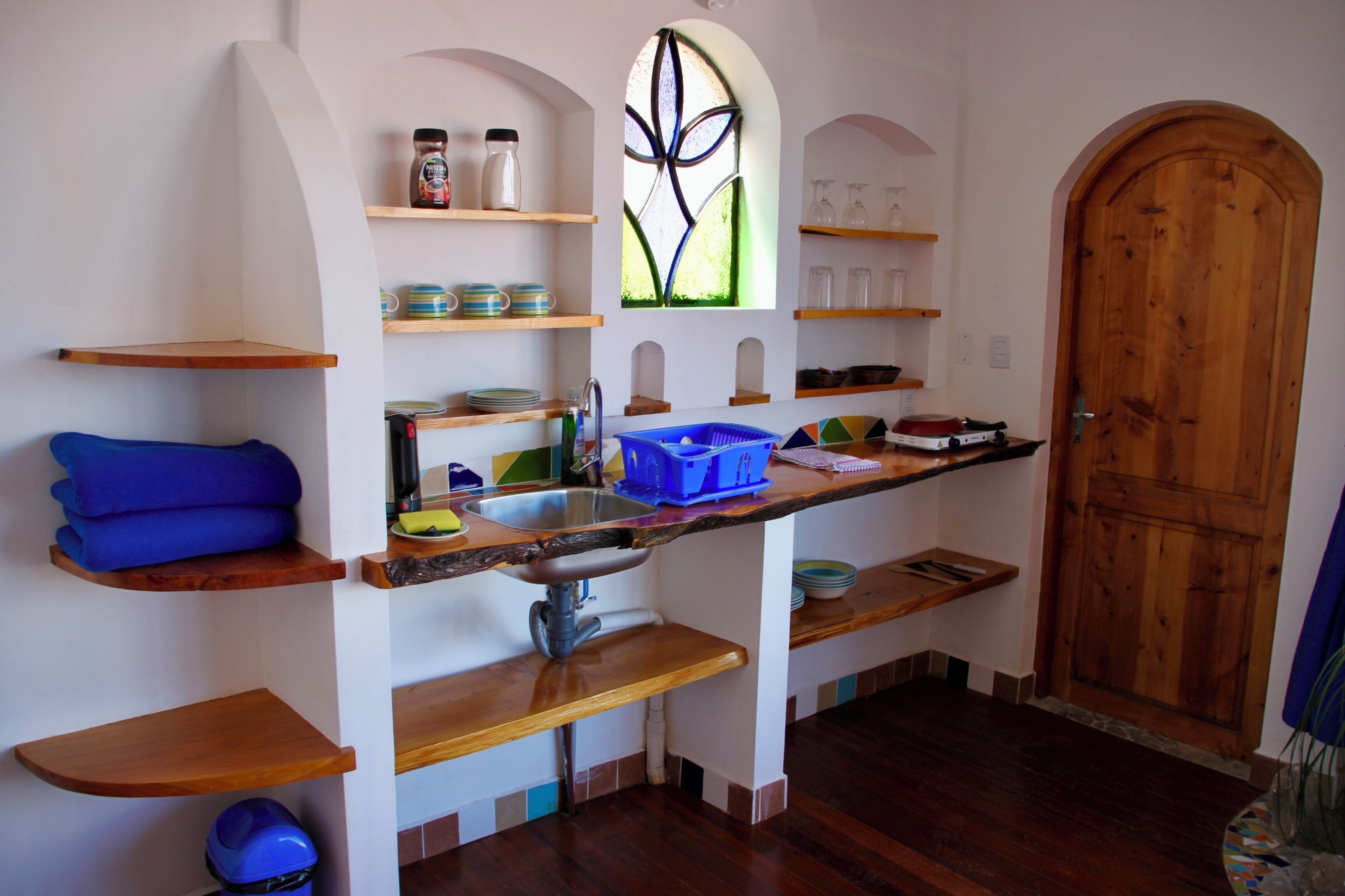 A compact kitchen with wooden shelves, a built-in sink, and a large window with a stained glass design.