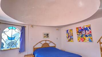 Ein Schlafzimmer mit einem Bett, einem Fenster mit blauer Vorhang und zwei farbenfrohen Bildern an der Wand.