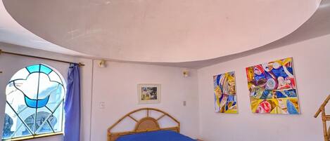 Un dormitorio con una cama, un ventanal con cortina azul y dos cuadros coloridos en la pared.