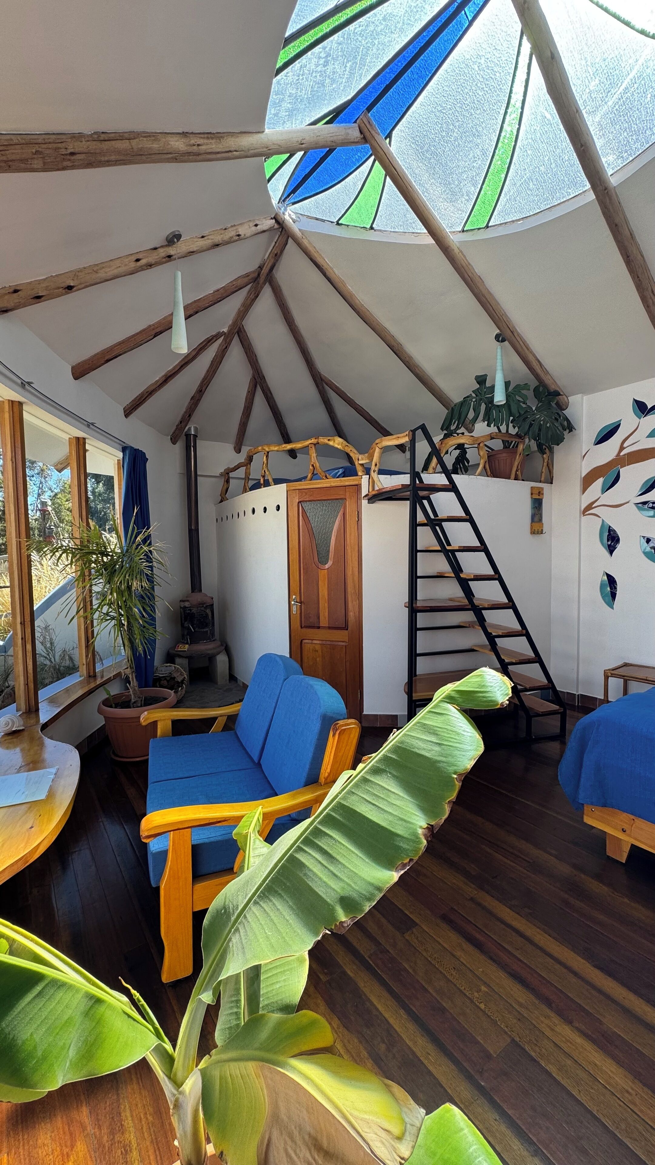 A cozy, rustic interior with a sloped ceiling, wooden beams, a blue sofa, a wooden ladder, and a large window.