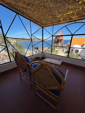 A room with a large window offering a view of the sea and buildings. There is a table and two wicker chairs. - Hotel La Cúpula (Copacabana)