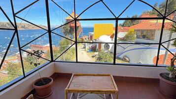 Ein Zimmer mit einem großen Fenster, das Meer und Gebäude zeigt. Es gibt einen kleinen Tisch und zwei Stühle mit blauen Kissen.