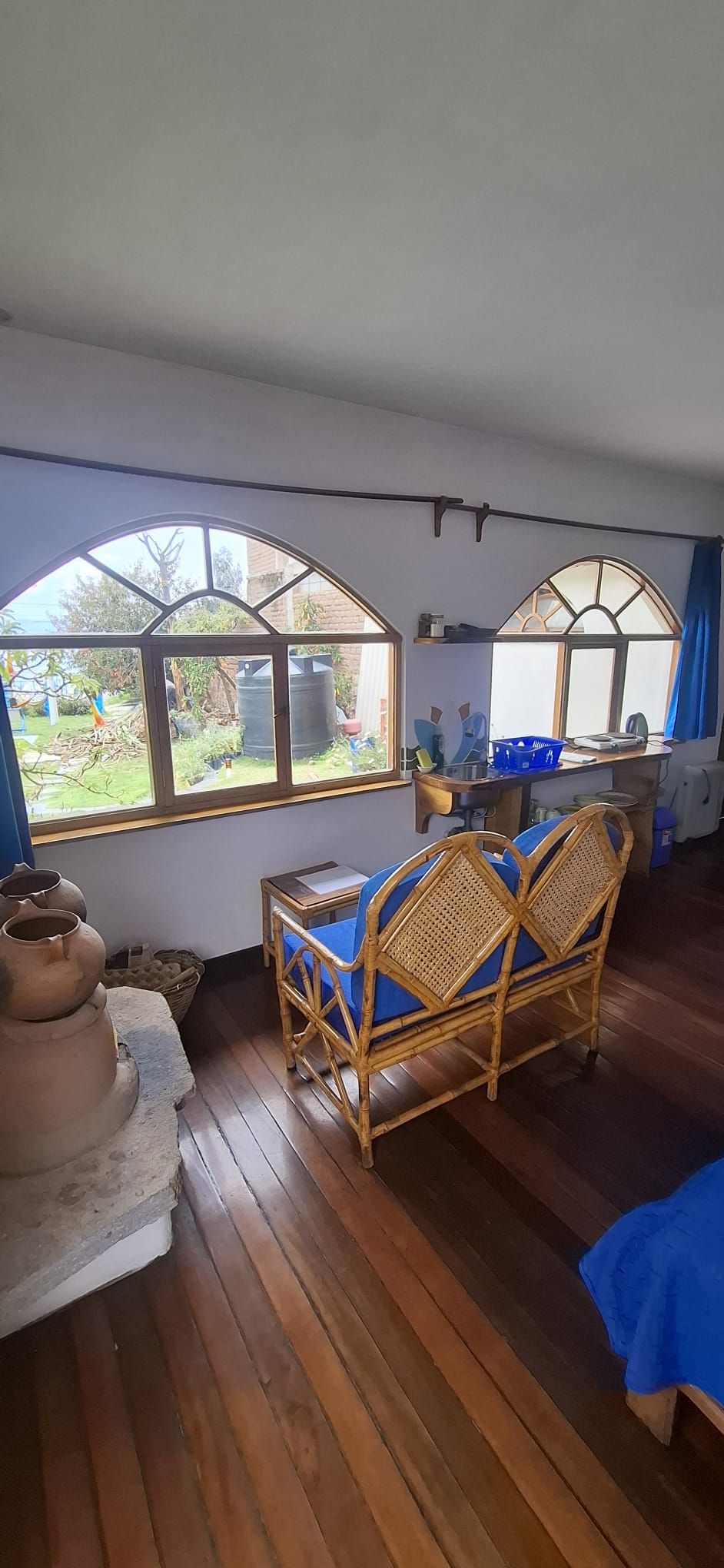 A room with wooden flooring, two wicker chairs with blue cushions, a table with a blue tablecloth, and large arched windows overlooking a garden.