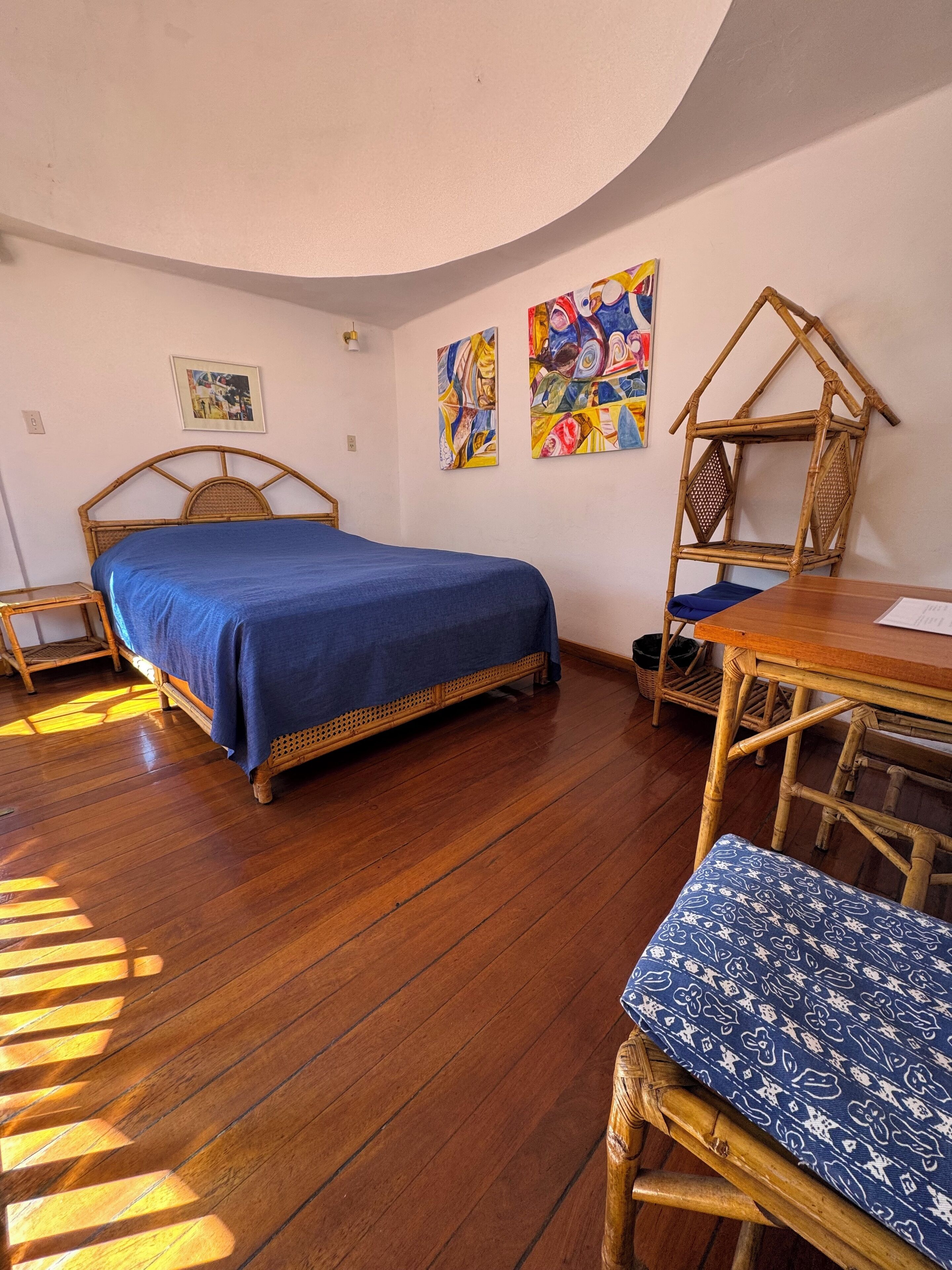 A room with a bed, a wooden table, a chair, and two wall-mounted paintings.