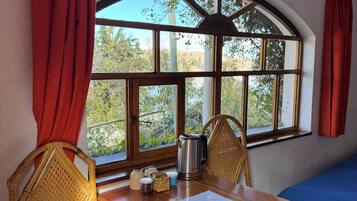 Una habitación con una mesa de madera, dos sillas de mimbre, un ventanal con cortinas rojas y vistas al verdor exterior.