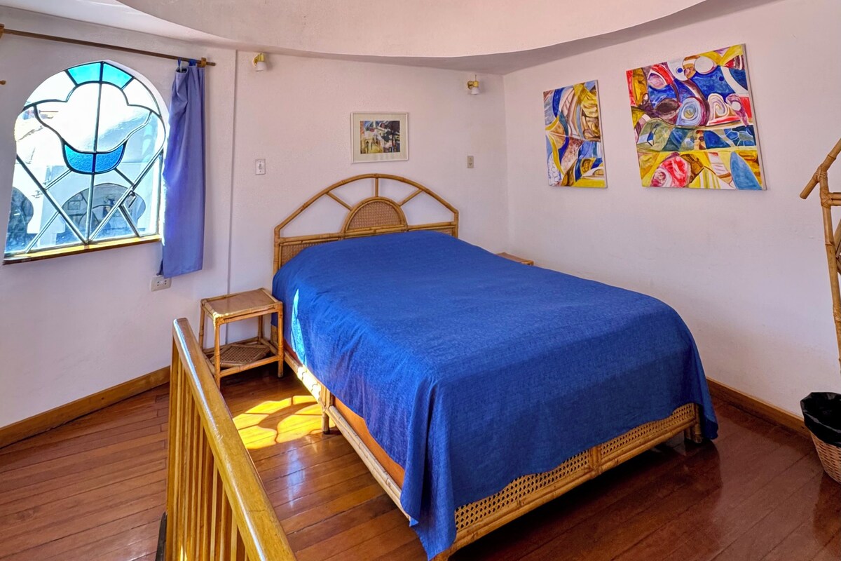 A bedroom with a bed, a window with a blue curtain, and two colorful paintings on the wall.