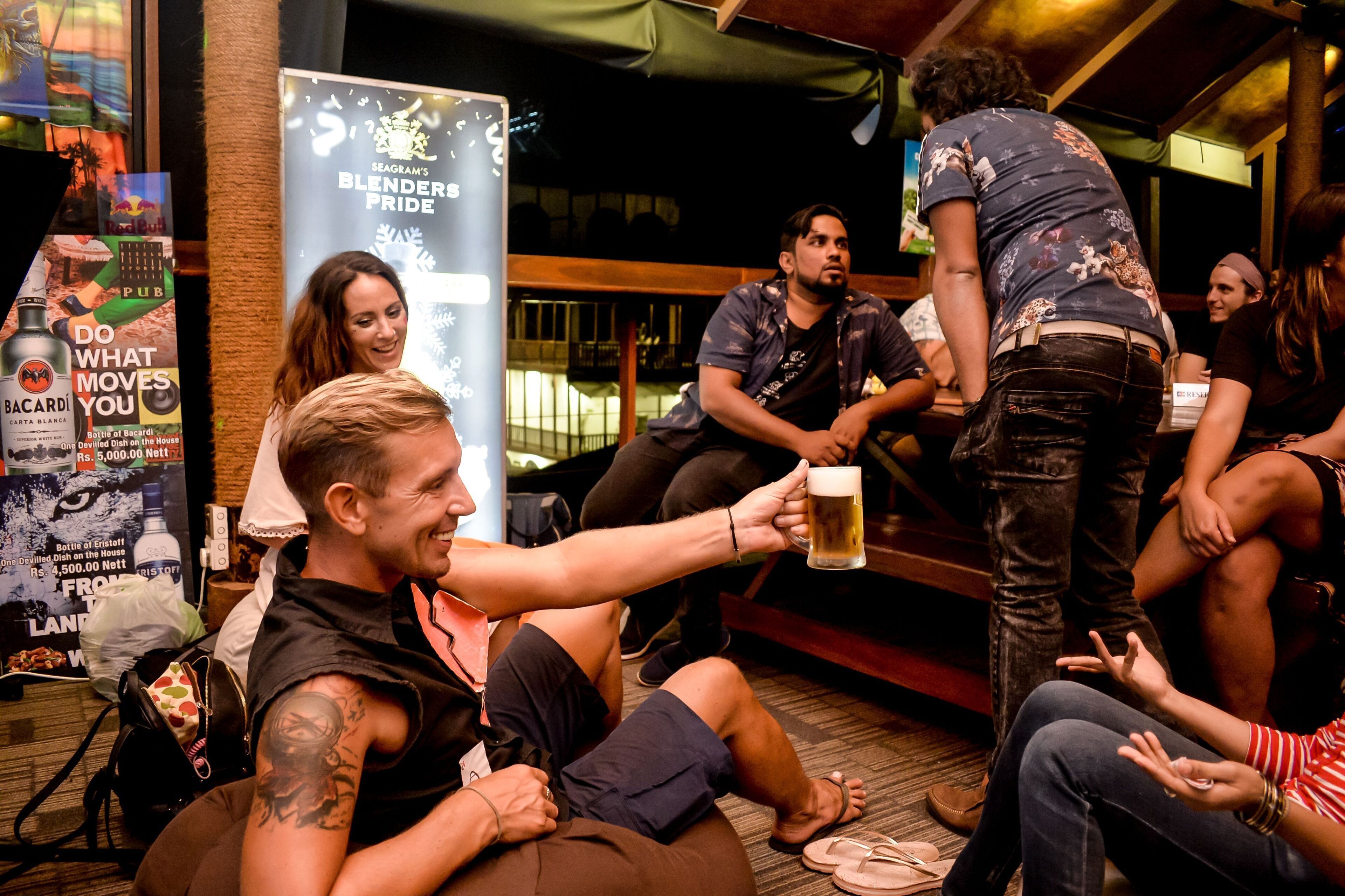 A group of people sitting and standing in a social setting, possibly a bar or lounge, with a poster advertising a beer brand in the background.