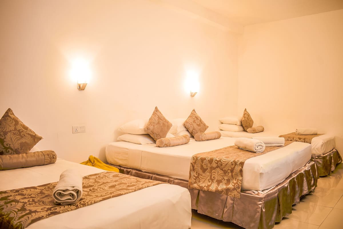 A hotel room with three beds, each with patterned bedspreads and pillows. There are wall-mounted lights and a visible electrical outlet.