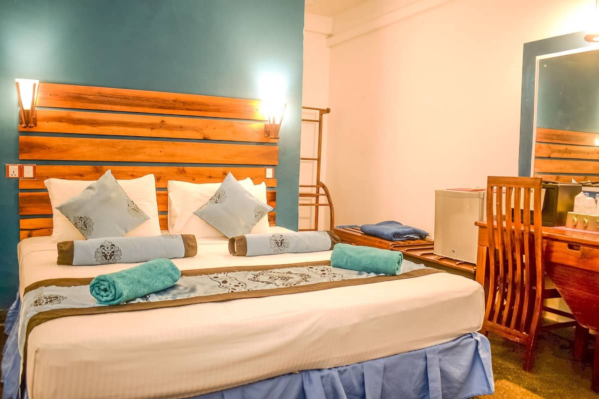 A hotel room with a wooden headboard, a bed with white bedding and patterned pillows, a blue skirted bedspread, and a wooden desk with a chair.