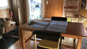 Une table de salle à manger en bois, accompagnée de deux chaises noires, d’un panier en osier et d’un extincteur placé près de la fenêtre.