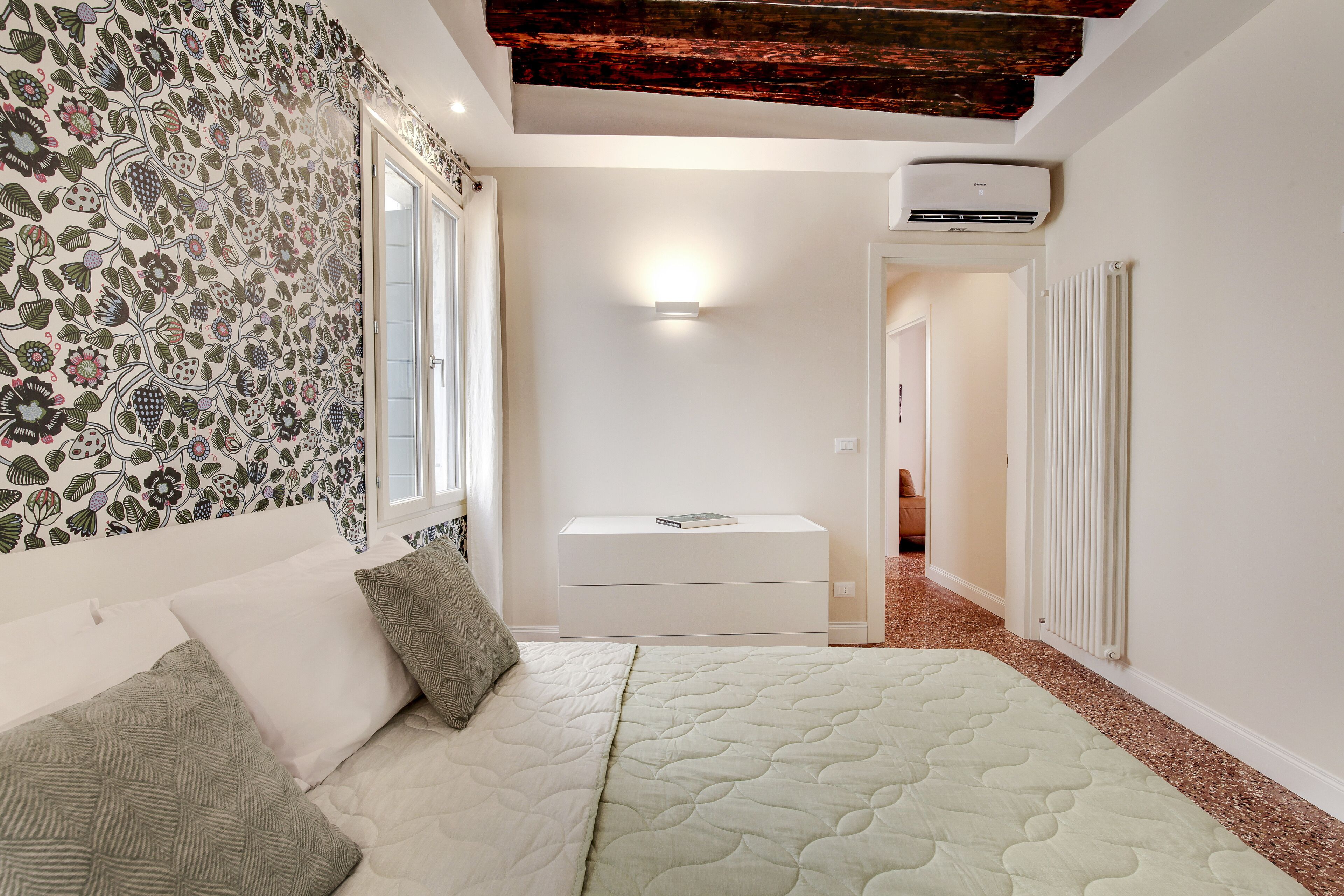 A bedroom with a bed, a nightstand, a wall-mounted air conditioner, and a window with curtains.