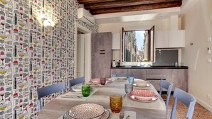 Una zona de menjador amb una taula rodona preparada per a sis persones, envoltada de cadires. L'espai destaca per un fons de paper pintat, una cuina moderna i una finestra amb vista a edificis.