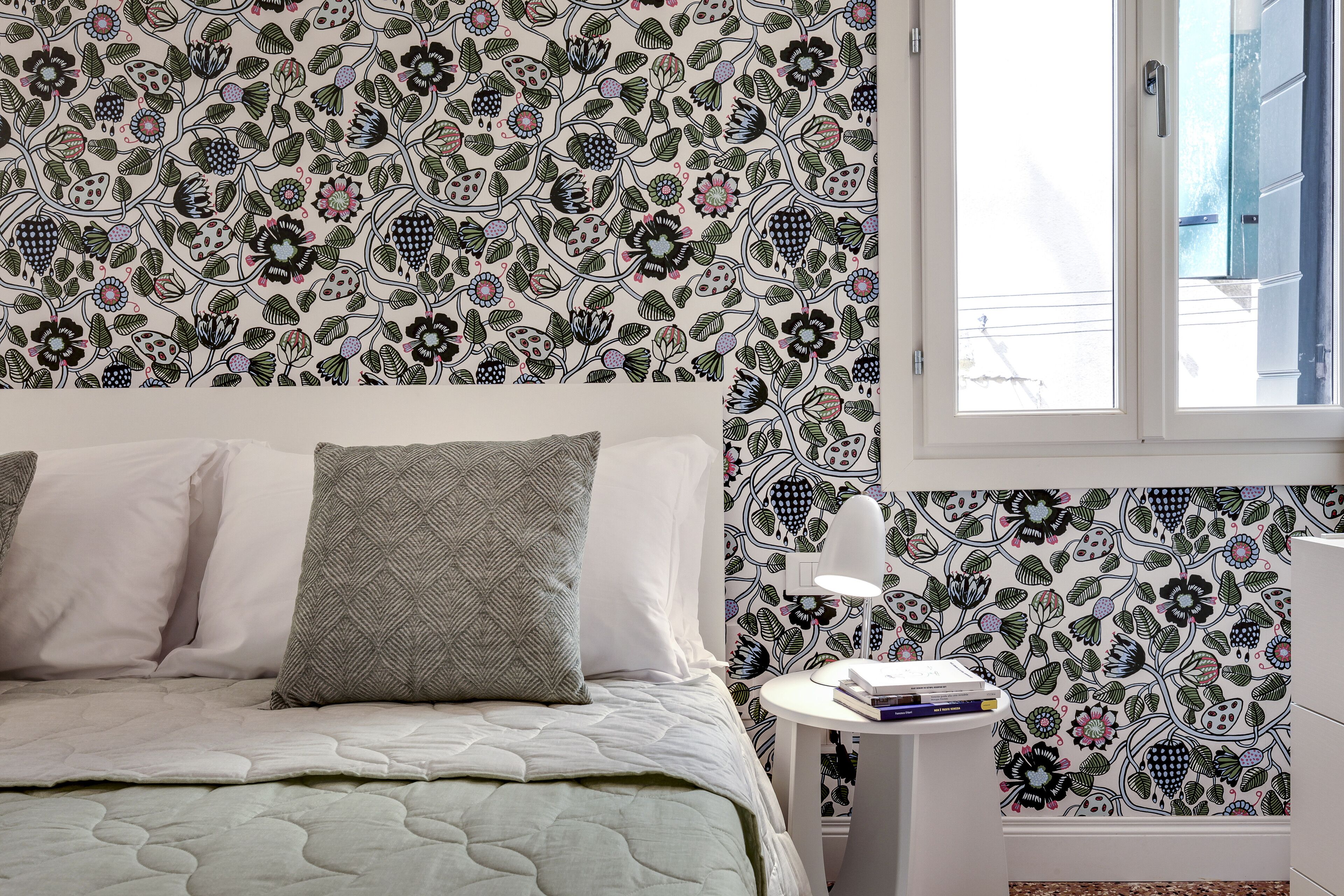 Una camera da letto con carta da parati decorata, un letto con cuscini, un comodino con una lampada e una finestra.