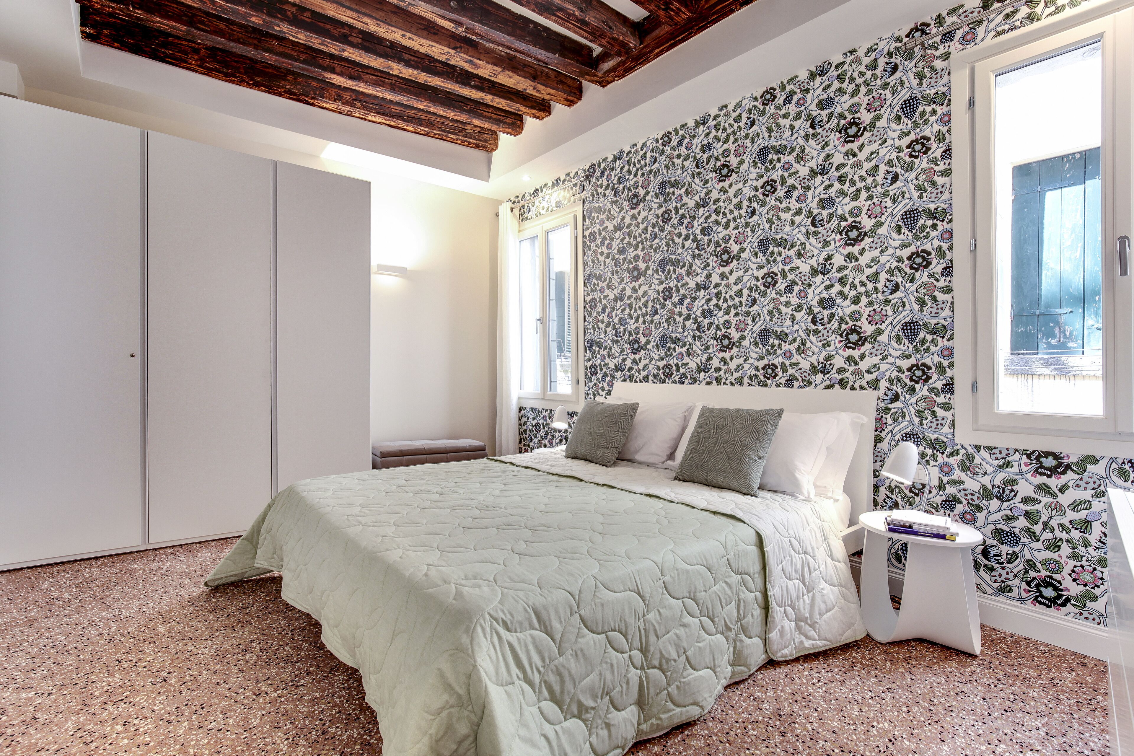 A bedroom with a bed, a nightstand, a wardrobe, and a window with patterned wallpaper.