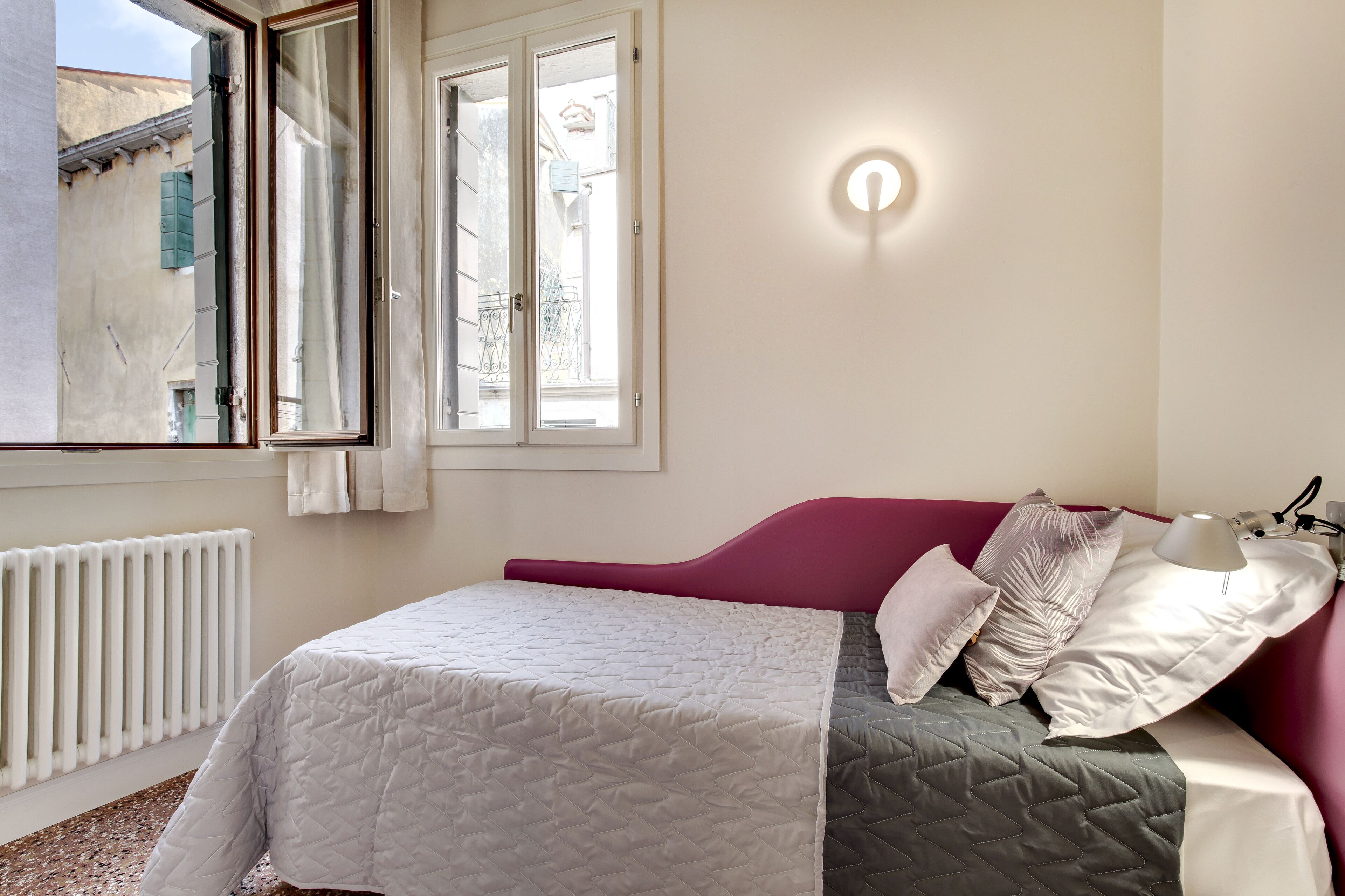 A bedroom with a bed, a window, a radiator, and a wall-mounted light fixture.