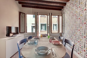 A dining area with a long table set for four, flanked by chairs, with a television on a white cabinet to the left.