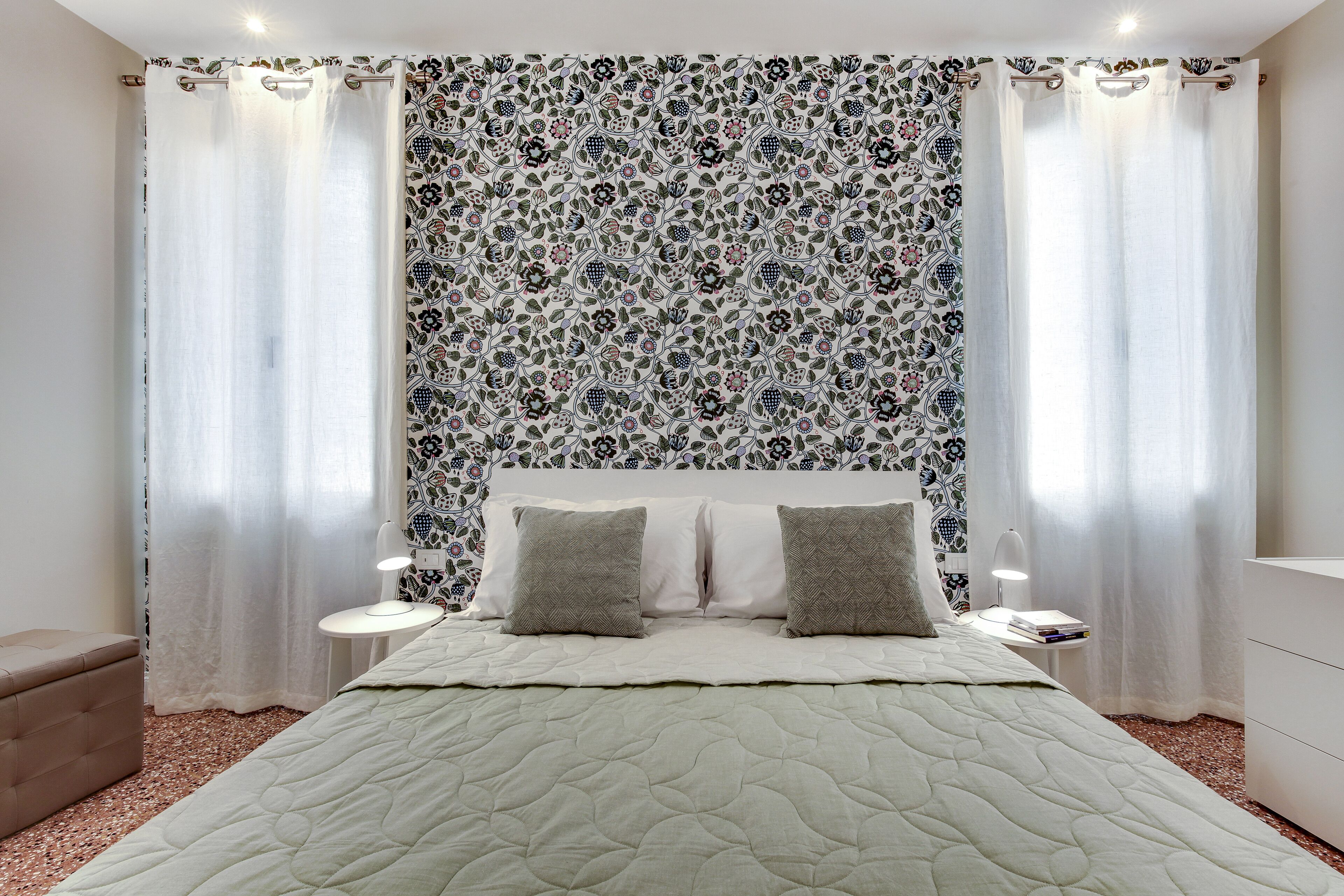 A bedroom with a bed, two pillows, a nightstand, a lamp, and a patterned wallpaper.