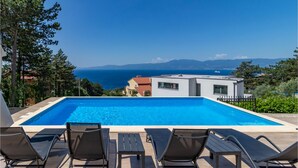 A swimming pool with lounge chairs, a view of the sea, and modern buildings in the background. - Beach front home in Kostrena (Kostrena)