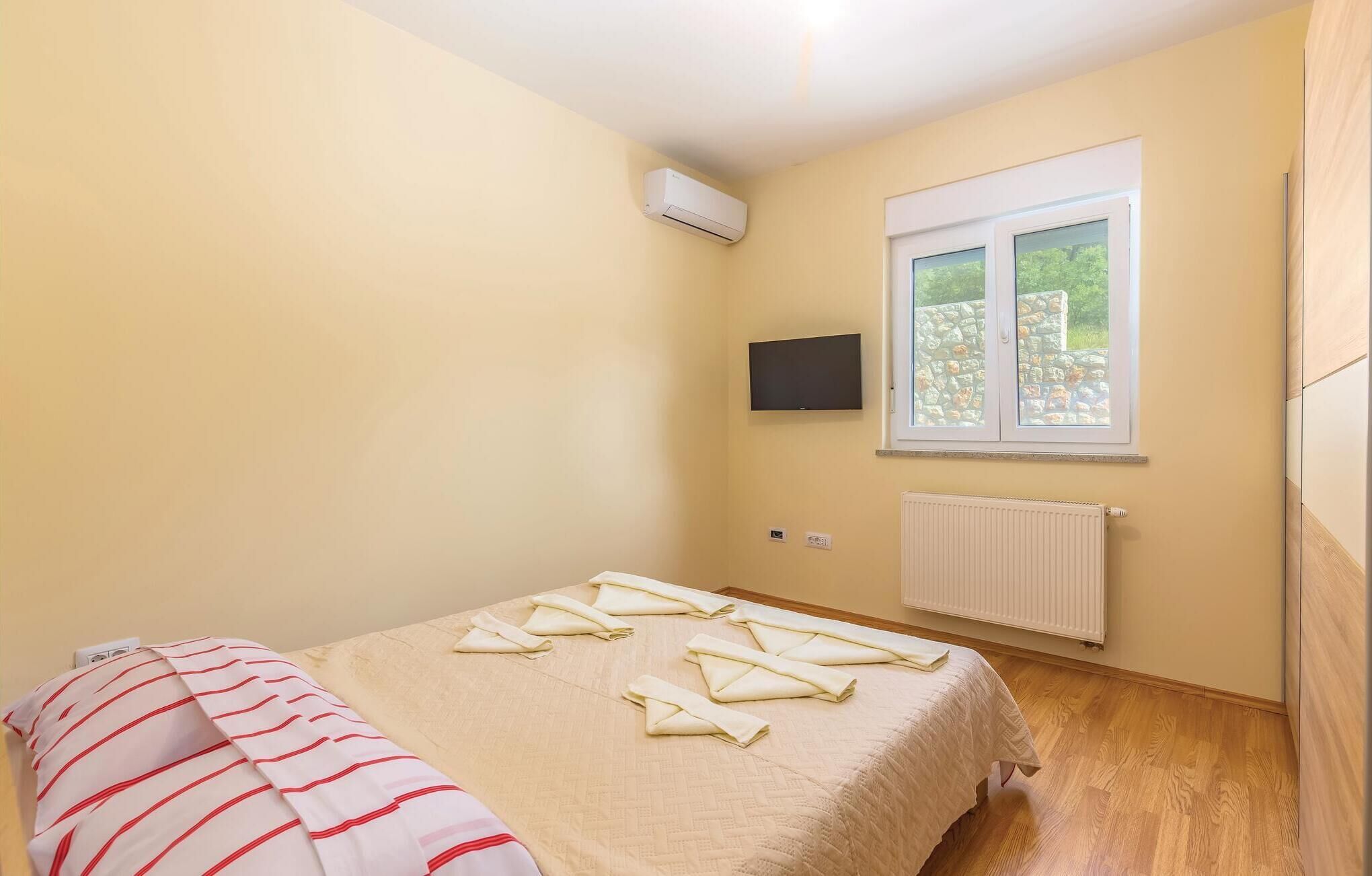 A small, well-lit room with a bed, a flat-screen TV mounted above a window, and a radiator below the window.