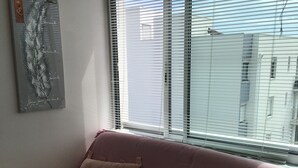 A pink sofa with two cushions, a framed artwork on the wall, and a window with blinds. - Sea view apartment (Quiberon)