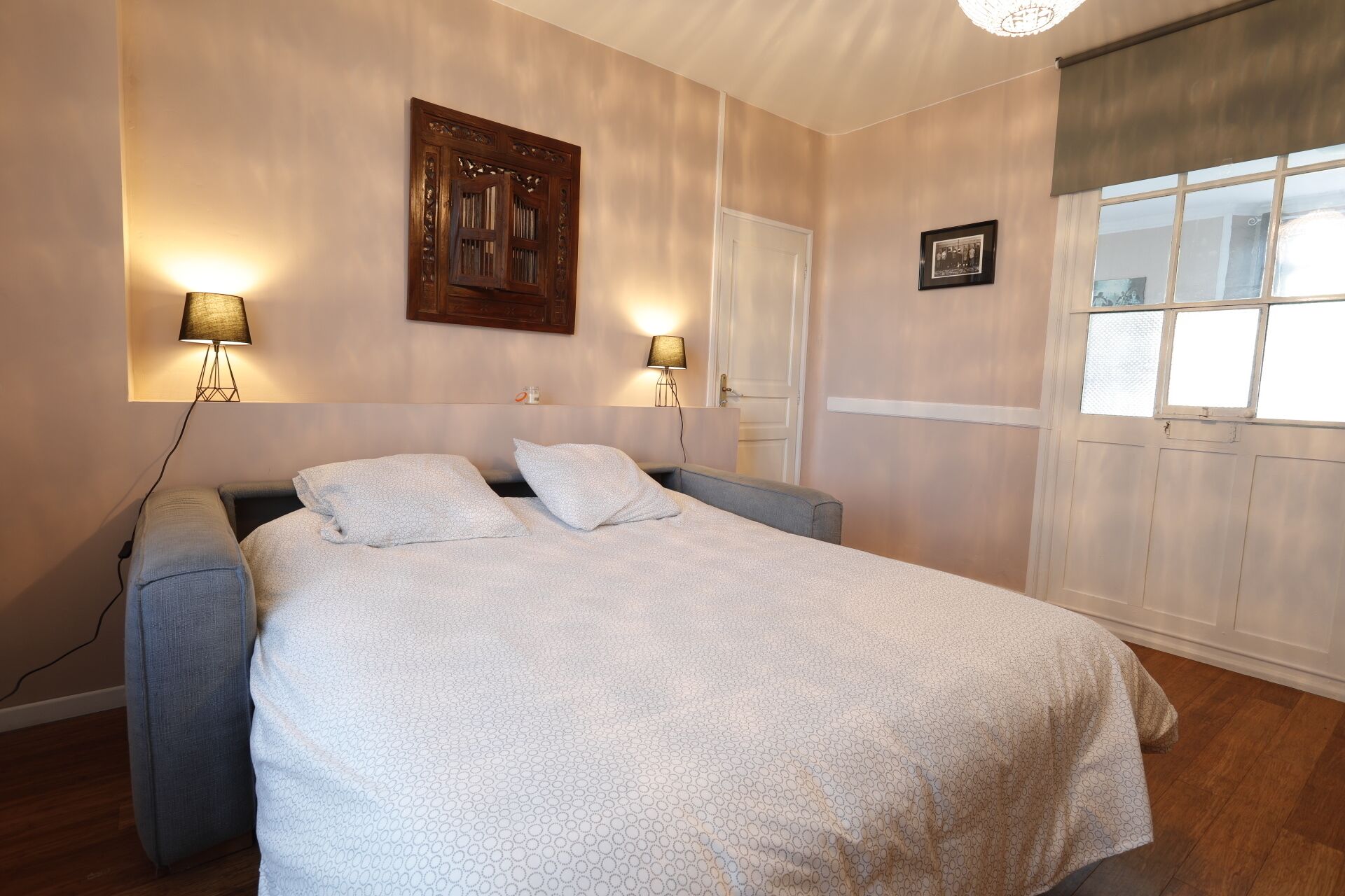 A bedroom with a bed, two bedside lamps, a wooden headboard, a door, and a window with blinds.