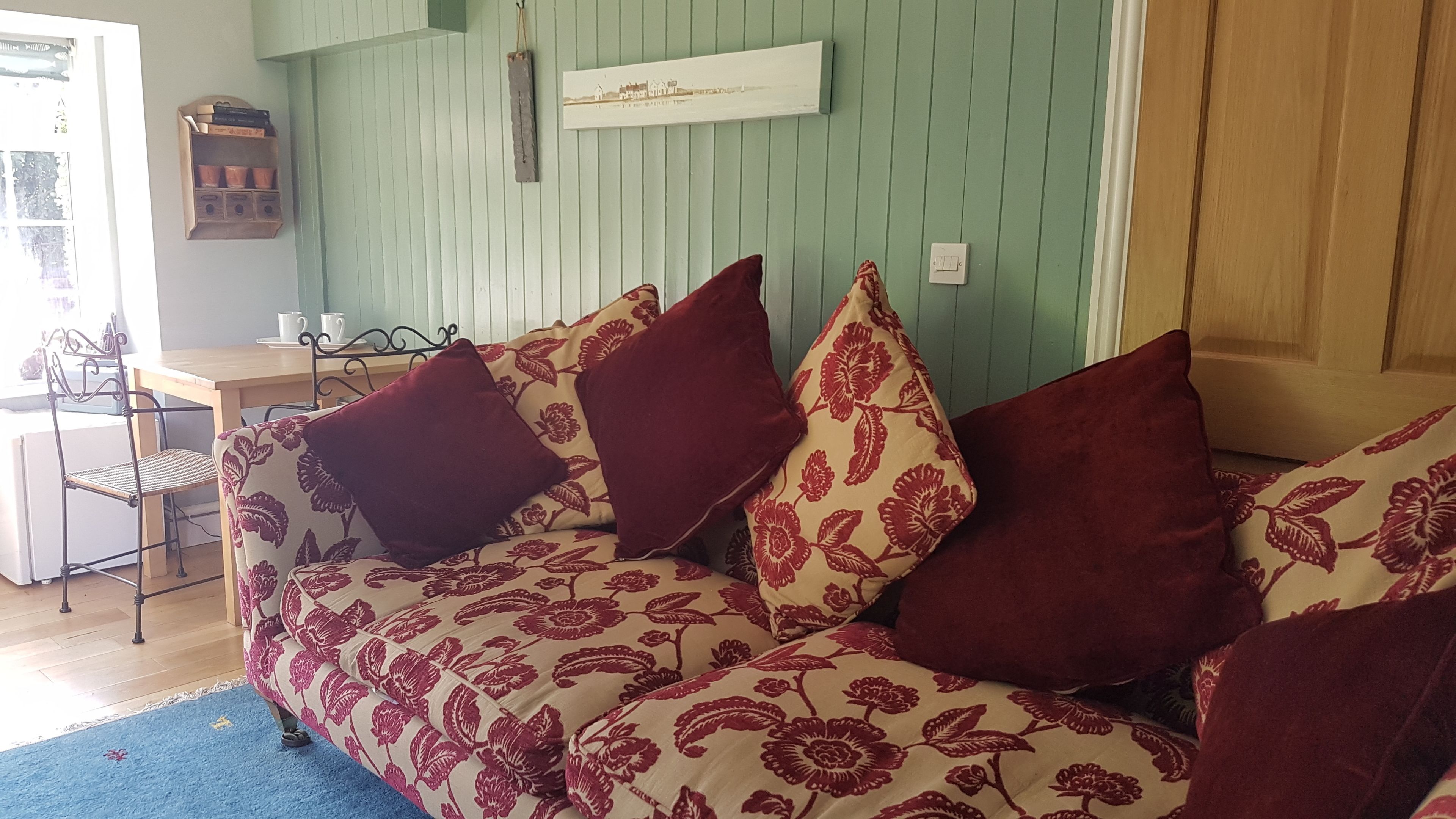 Une pièce chaleureuse avec un canapé agrémenté de coussins à motifs floraux, une chaise en bois, une petite table et une étagère fixée au mur.