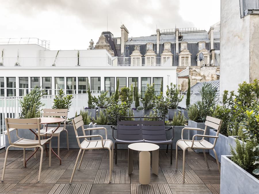 Terraza en la azotea con mobiliario exterior, incluyendo sillas y una mesa, rodeada de macetas con plantas y vistas a un paisaje urbano.
