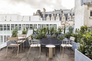 A rooftop terrace with outdoor seating, including chairs and a table, surrounded by potted plants and overlooking a cityscape. - Le Grand Quartier (Paris)