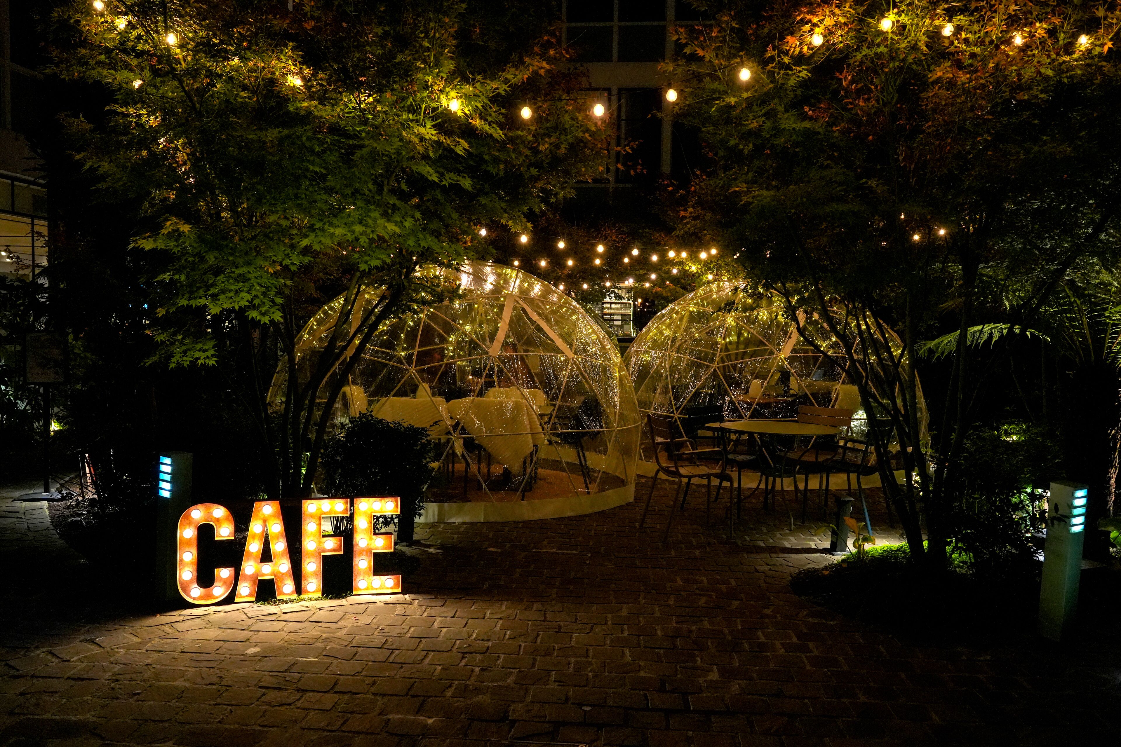 Ένα καφέ με μια φωτεινή επιγραφή που γράφει «CAFE», τραπέζια σε εξωτερικό χώρο κάτω από διαφανείς ομπρέλες και γιρλάντες με φωτάκια.