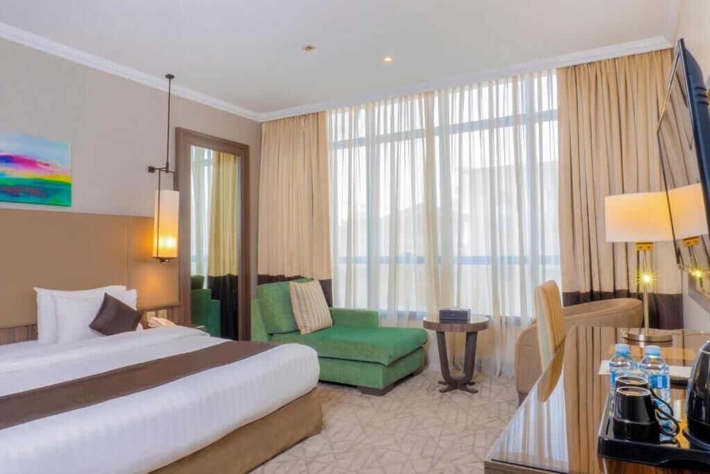 Habitación de hotel con una cama grande, un sillón verde, una mesita de noche con lámpara, y un escritorio con silla y cafetera.