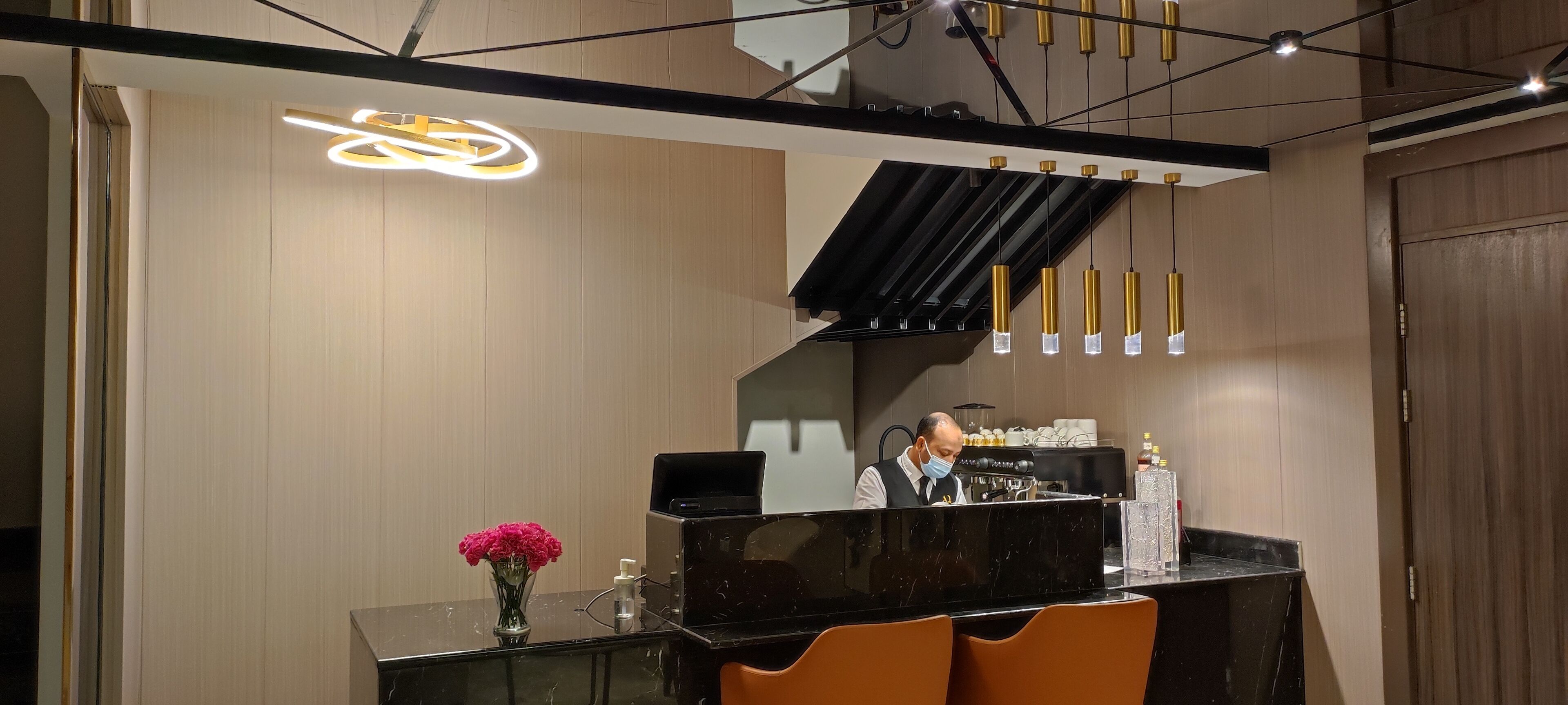Een moderne bar met een zwarte marmeren toonbank, oranje stoelen, een barista en een bijzondere plafondlamp.