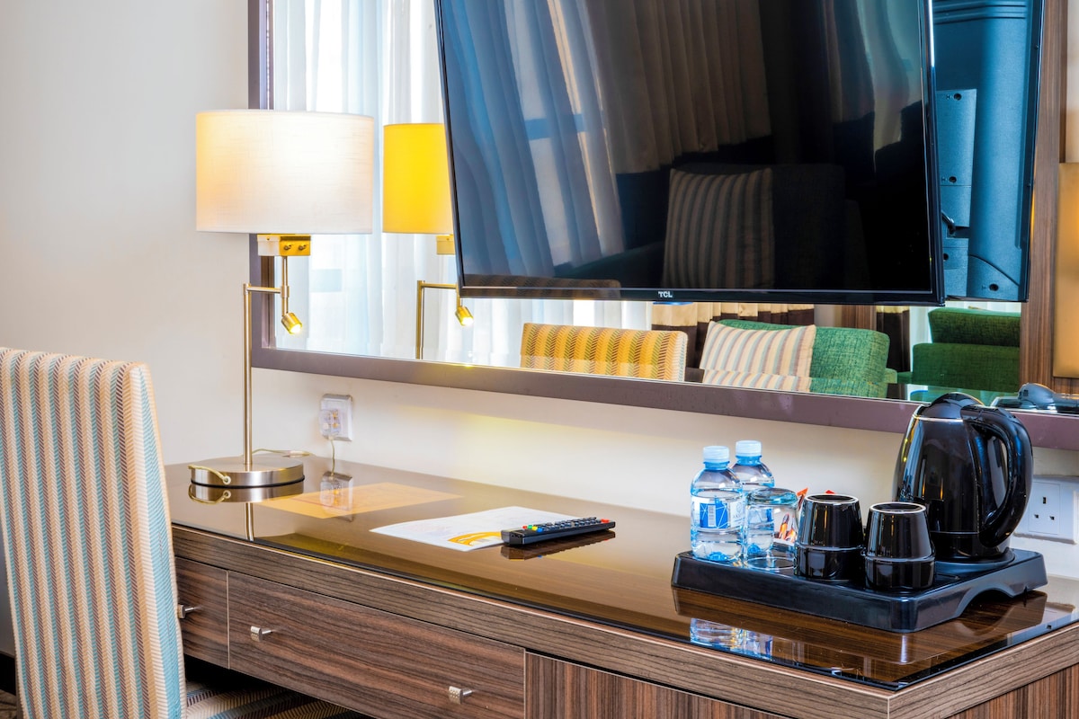 A hotel room with a wooden desk, a flat-screen TV, a mirror, a lamp, a remote control, a tray with water bottles, cups, and a coffee maker.