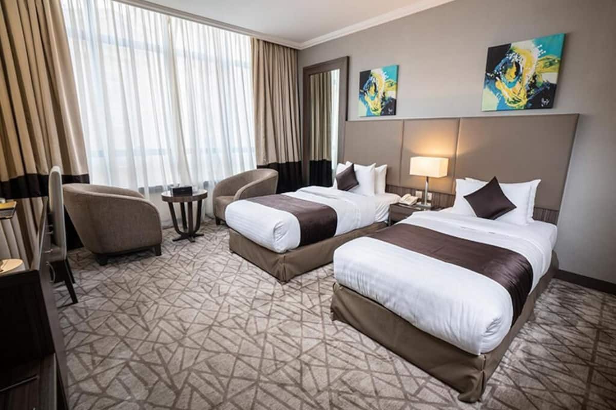 Une chambre d’hôtel avec deux lits, un bureau, une chaise, une petite table et une grande fenêtre avec des rideaux.