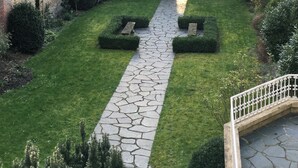Une cour avec un chemin en pierre, des haies taillées et un mur en briques.