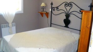 A neatly made bed in a room with a metal headboard, a small table with a lamp, and a window with curtains. - Les Mazets Du Soleil: Bed and Breakfast (Maussane-les-Alpilles)