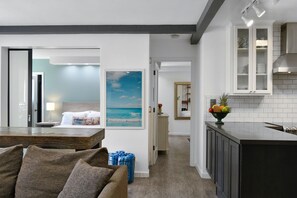 Relax in a serene space with a large bed, a dining table, and a sofa, surrounded by a coastal-inspired artwork and a well-appointed kitchen.