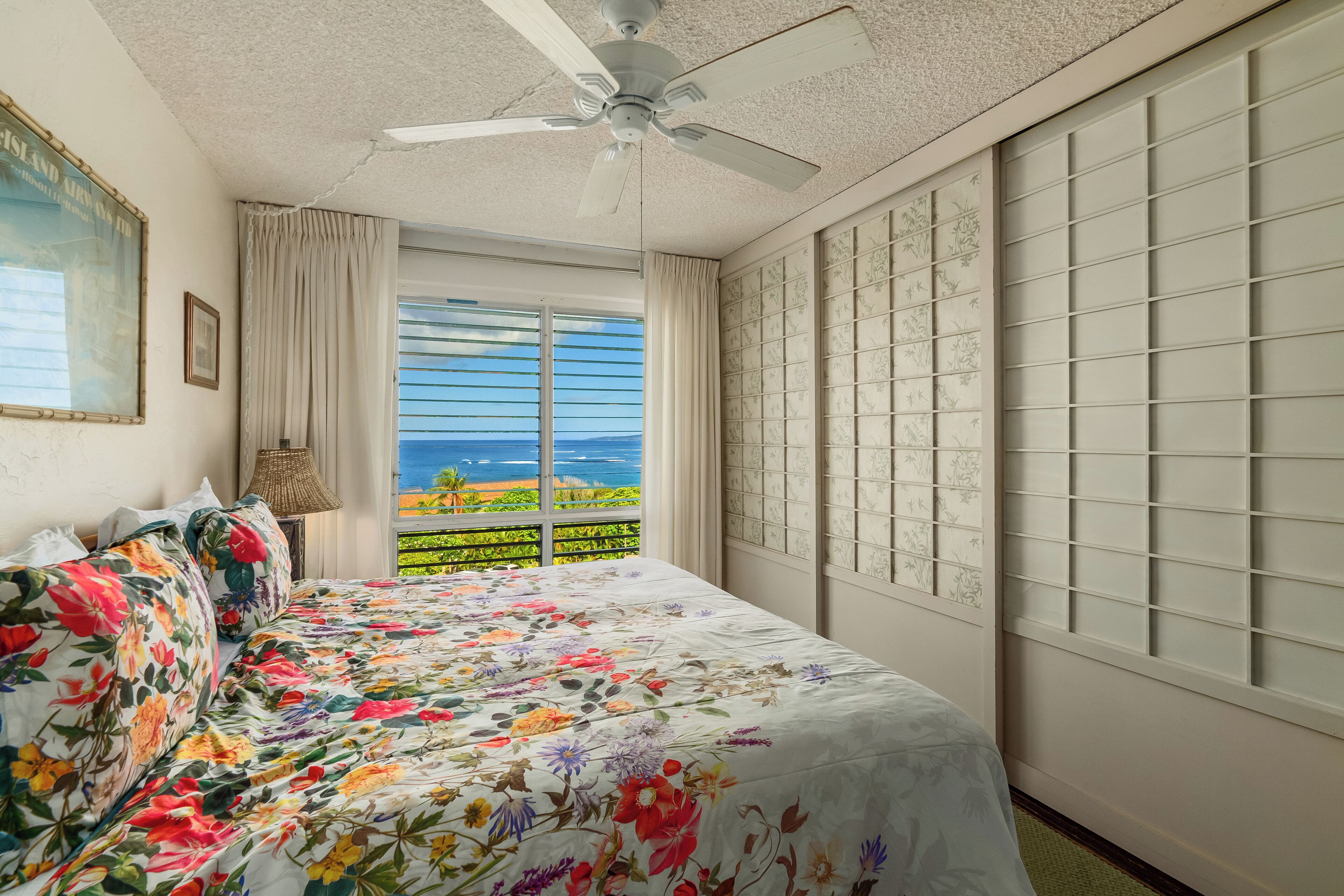A bedroom with a bed, a ceiling fan, a window with a view of the ocean, and a decorative wall panel.