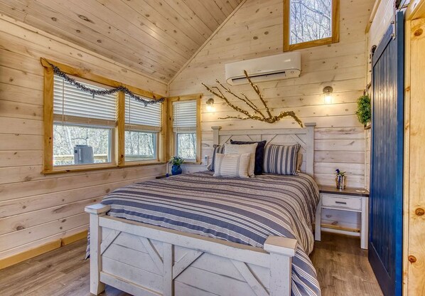 Un dormitorio de cabaña de madera con cama, mesitas de noche y ventana con persianas.