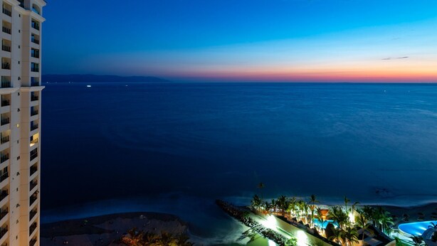 Un resort con vista al océano al anochecer, que cuenta con una zona frente a la playa con palmeras y una piscina.