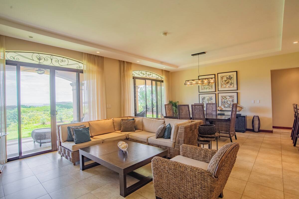 A spacious living room with a large sofa, a coffee table, and wicker chairs. There is a dining area with a table and chairs. Large windows provide a view of the outdoors.